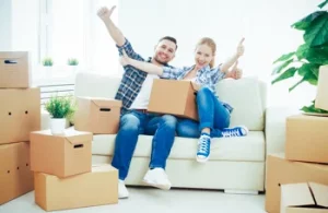 Selfstorage Erfahrungen - Zwei Personen sitzen auf einer Bank inmitten von Umzugskartons und zeigen mit dem Daumen hoch.